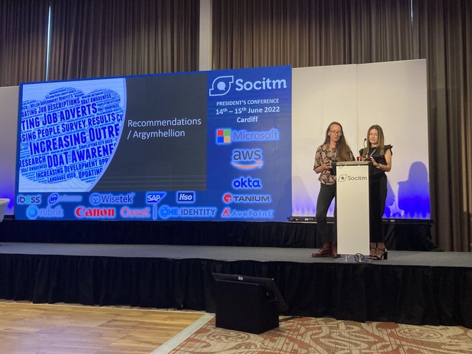Two women standing at a podium on a stage. Behind them is a massive screen that's displaying their presentation slides.