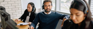 A man wearing headphones, sitting by an office desk, looking into the camera, two women speaking on the phone next to him