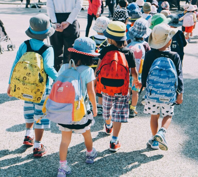 A small group of small children) seen from behind) all wearing school packpacks.