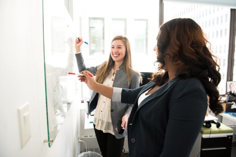 Two women writing on a dry-erase glass wall board