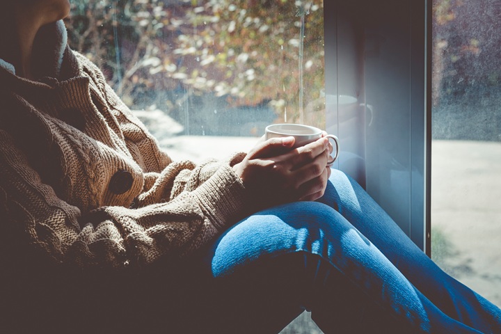 Woman drinking a cup of tea, sitting in a window seat with autumnal colours outside.