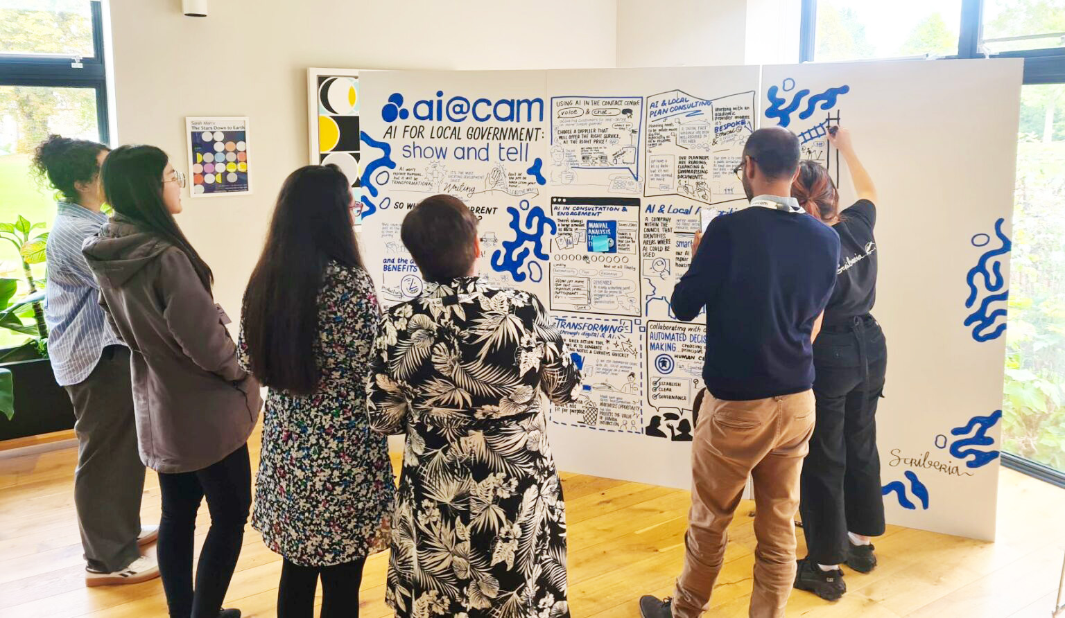 Group of people standing in front of a large banner full of illustrations and words around ai@cam: AI for local government