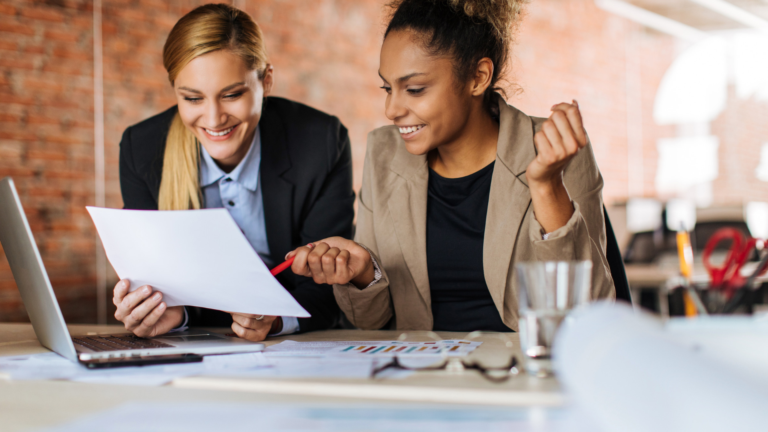 2 women sitting side-by-side at a desk looking at a piece of paper instead of the laptop in front of them both.